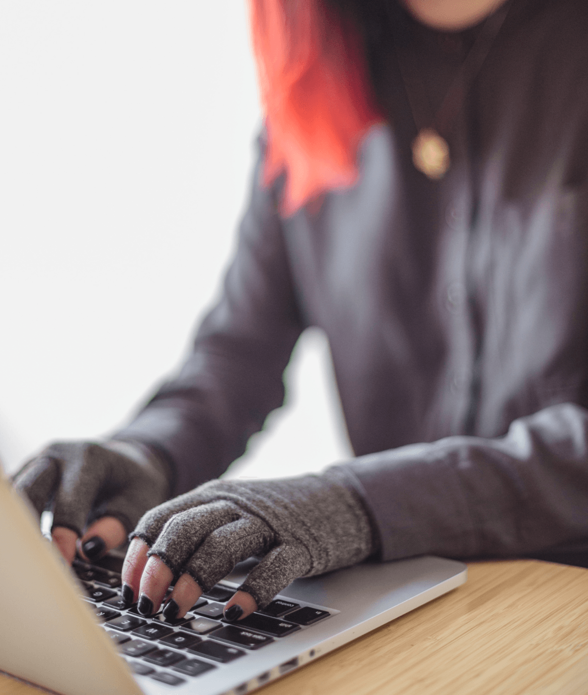 Uma pessoa asiática com deficiência e de gênero fluido está digitando em um laptop enquanto usa luvas de compressão. As mãos e o teclado são o ponto focal.
