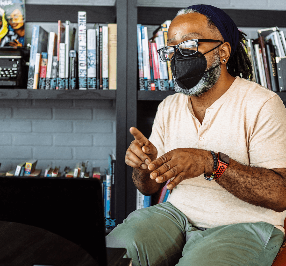 Retrato de um homem negro surdo, usando óculos e máscara KN-95, fazendo uma chamada de vídeo em linguagem de sinais utilizando um laptop. Um homem sentado em uma mesa da lanchonete de uma biblioteca comunitária repleta de livros e pôsteres.