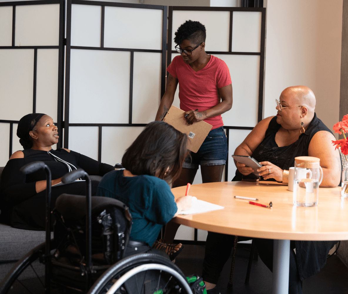 Quatro pessoas não brancas, portadoras de deficiências, se reúnem em torno de uma mesa durante uma reunião. Uma mulher negra sentada em um sofá fala com uma expressão neutra enquanto as outras três pessoas (uma pessoa do sul da Ásia sentada em uma cadeira de rodas e fazendo anotações, uma pessoa negra não-binária sentada em uma cadeira com um tablet e uma bengala, e uma pessoa negra não-binária de pé com uma prancheta) ouvem atentamente.